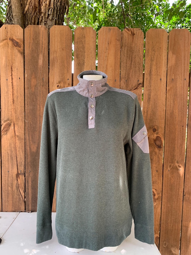 May include: A green long-sleeved sweater with a mock neck and button closure. The sweater has a gray patch on the shoulder and a gray patch on the left sleeve.