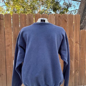 May include: A navy blue crew neck sweatshirt. The long-sleeved top is displayed on a mannequin against a wooden fence and a backdrop of trees. The sweatshirt is a solid color and appears to be made of a soft material.