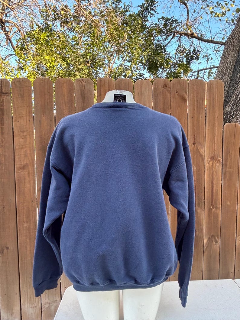 May include: A navy blue crew neck sweatshirt. The long-sleeved top is displayed on a mannequin against a wooden fence and a backdrop of trees. The sweatshirt is a solid color and appears to be made of a soft material.