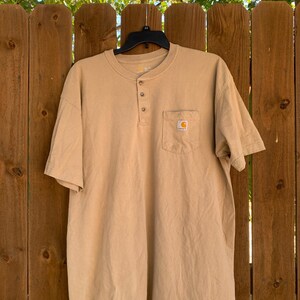 K&ouml;nnte beinhalten: Ein hellbraunes kurz&auml;rmeliges Henley-Shirt mit einer Brusttasche. Das Shirt hat drei Kn&ouml;pfe am Halsausschnitt und ein kleines Logo auf der Tasche. Das Shirt h&auml;ngt an einem schwarzen Kleiderb&uuml;gel vor einem Holzzaun.