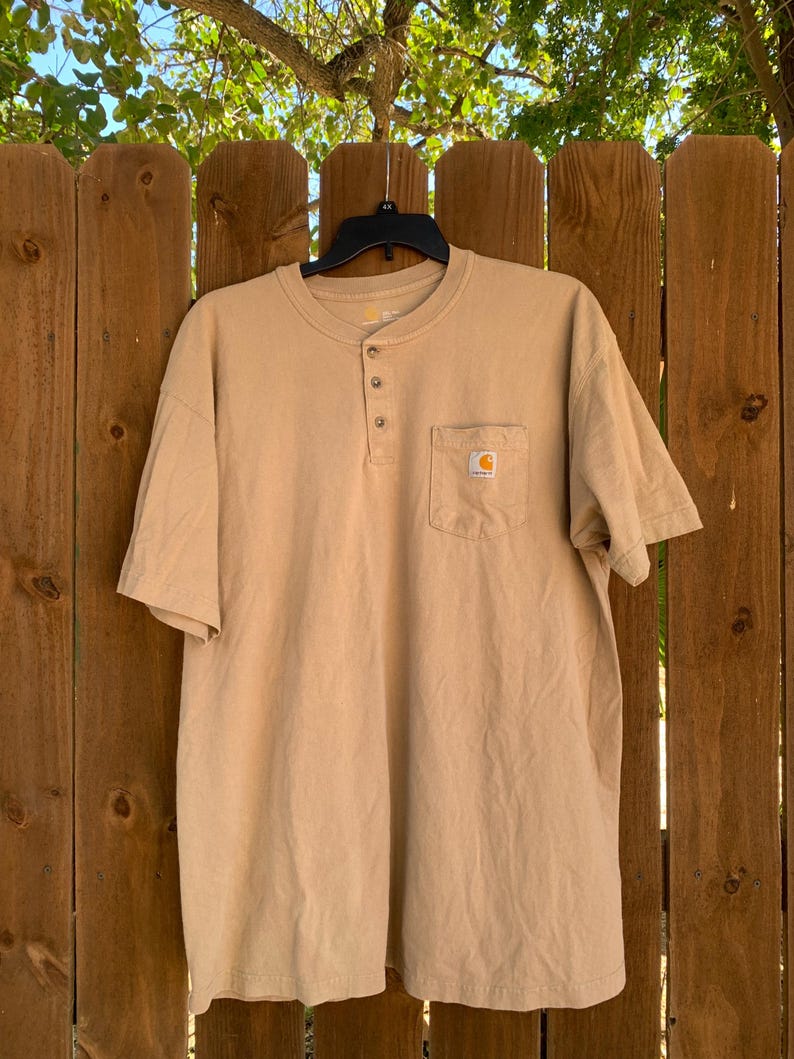 K&ouml;nnte beinhalten: Ein hellbraunes kurz&auml;rmeliges Henley-Shirt mit einer Brusttasche. Das Shirt hat drei Kn&ouml;pfe am Halsausschnitt und ein kleines Logo auf der Tasche. Das Shirt h&auml;ngt an einem schwarzen Kleiderb&uuml;gel vor einem Holzzaun.