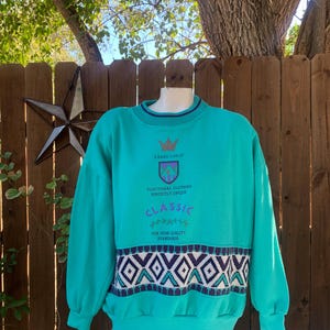 Puede incluir: Sudadera de color turquesa con cuello redondo y mangas largas. La sudadera presenta un estampado geom&eacute;trico en blanco, morado y turquesa en la parte inferior. El texto de la sudadera dice: "A BASIC LINE OF FUNCTIONAL CLOTHING PERFECTLY UNIQUE CLASSIC FOR HIGH QUALITY STANDARDS."
