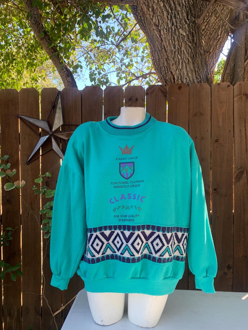 Puede incluir: Sudadera de color turquesa con cuello redondo y mangas largas. La sudadera presenta un estampado geom&eacute;trico en blanco, morado y turquesa en la parte inferior. El texto de la sudadera dice: "A BASIC LINE OF FUNCTIONAL CLOTHING PERFECTLY UNIQUE CLASSIC FOR HIGH QUALITY STANDARDS."