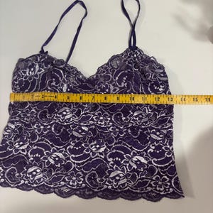 May include: A dark purple camisole with white floral lace detailing. The camisole has thin, adjustable straps and a scalloped hem. The garment measures approximately 16 inches across. The fabric appears to be delicate and semi-sheer.