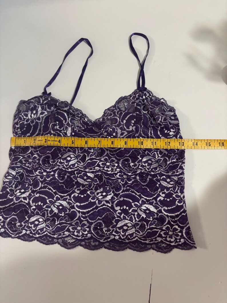 May include: A dark purple camisole with white floral lace detailing. The camisole has thin, adjustable straps and a scalloped hem. The garment measures approximately 16 inches across. The fabric appears to be delicate and semi-sheer.