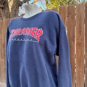 May include: Navy blue sweatshirt with the text "THRASHER MAGAZINE" in red and white. The long-sleeved crew neck sweatshirt is displayed on a mannequin. The background includes a wooden fence and tree branches.