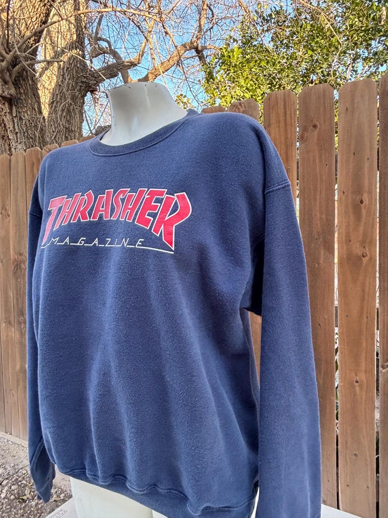 May include: Navy blue sweatshirt with the text "THRASHER MAGAZINE" in red and white. The long-sleeved crew neck sweatshirt is displayed on a mannequin. The background includes a wooden fence and tree branches.