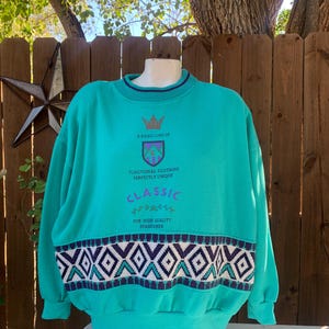 Puede incluir: Sudadera de color turquesa con cuello redondo y mangas largas. El dise&ntilde;o incluye un patr&oacute;n geom&eacute;trico en blanco, azul marino y morado en la parte inferior, y el texto "A Basic Line of Classic Functional Clothing Perfectly Unique".