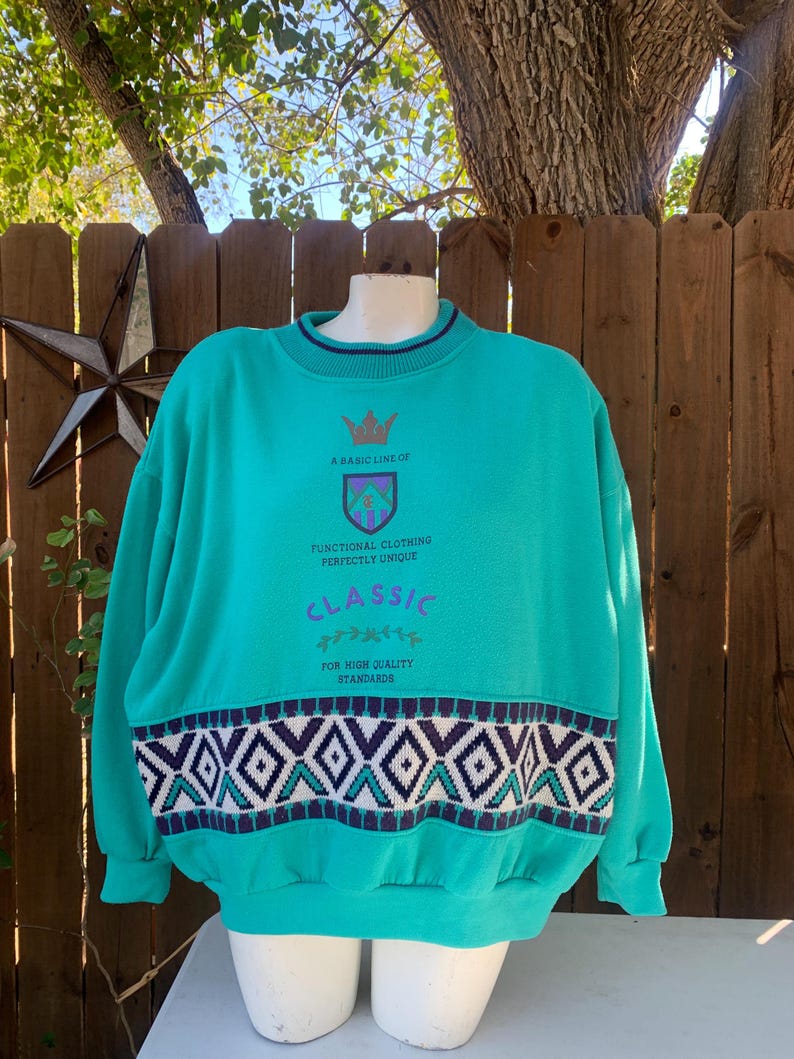 Puede incluir: Sudadera de color turquesa con cuello redondo y mangas largas. El dise&ntilde;o incluye un patr&oacute;n geom&eacute;trico en blanco, azul marino y morado en la parte inferior, y el texto "A Basic Line of Classic Functional Clothing Perfectly Unique".