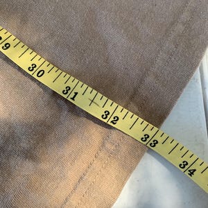 K&ouml;nnte beinhalten: Nahaufnahme eines beigefarbenen Stoffes mit einem gelben Ma&szlig;band. Das Band zeigt Messungen von 73,66 cm bis 86,36 cm. Der Stoff hat eine sichtbare Naht am Rand.