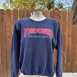 May include: Navy blue sweatshirt with the text "Thrasher Magazine" in red and white. The sweatshirt has a crew neck and long sleeves. The text is in a bold, stylized font.