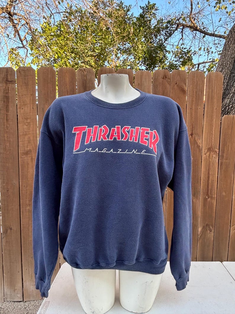 May include: Navy blue sweatshirt with the text "Thrasher Magazine" in red and white. The sweatshirt has a crew neck and long sleeves. The text is in a bold, stylized font.