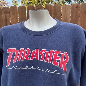 May include: Navy blue sweatshirt with the word "THRASHER" in red, outlined in white, arched across the chest. Below the word "THRASHER" is the text "MAGAZINE" in white. The sweatshirt is displayed on a mannequin.