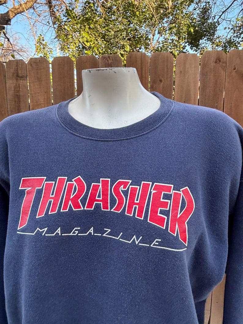 May include: Navy blue sweatshirt with the word "THRASHER" in red, outlined in white, arched across the chest. Below the word "THRASHER" is the text "MAGAZINE" in white. The sweatshirt is displayed on a mannequin.
