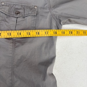 K&ouml;nnte beinhalten: Nahaufnahme eines grauen Hemdes mit einer Knopfleistentasche und einem Knopfverschluss. Das Hemd wird mit einem gelben Ma&szlig;band gemessen. Der &Auml;rmel des Hemdes wird von 40,6 cm bis 55,9 cm gemessen.