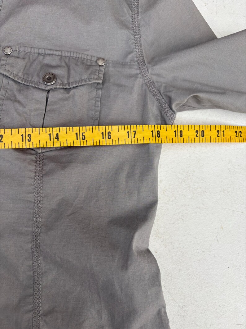 K&ouml;nnte beinhalten: Nahaufnahme eines grauen Hemdes mit einer Knopfleistentasche und einem Knopfverschluss. Das Hemd wird mit einem gelben Ma&szlig;band gemessen. Der &Auml;rmel des Hemdes wird von 40,6 cm bis 55,9 cm gemessen.