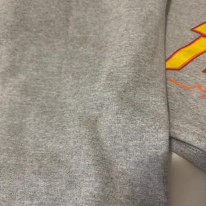 May include: A close-up of a gray sweatshirt. The sleeve is visible, with a design in yellow and orange on the upper right. The fabric has some discoloration.
