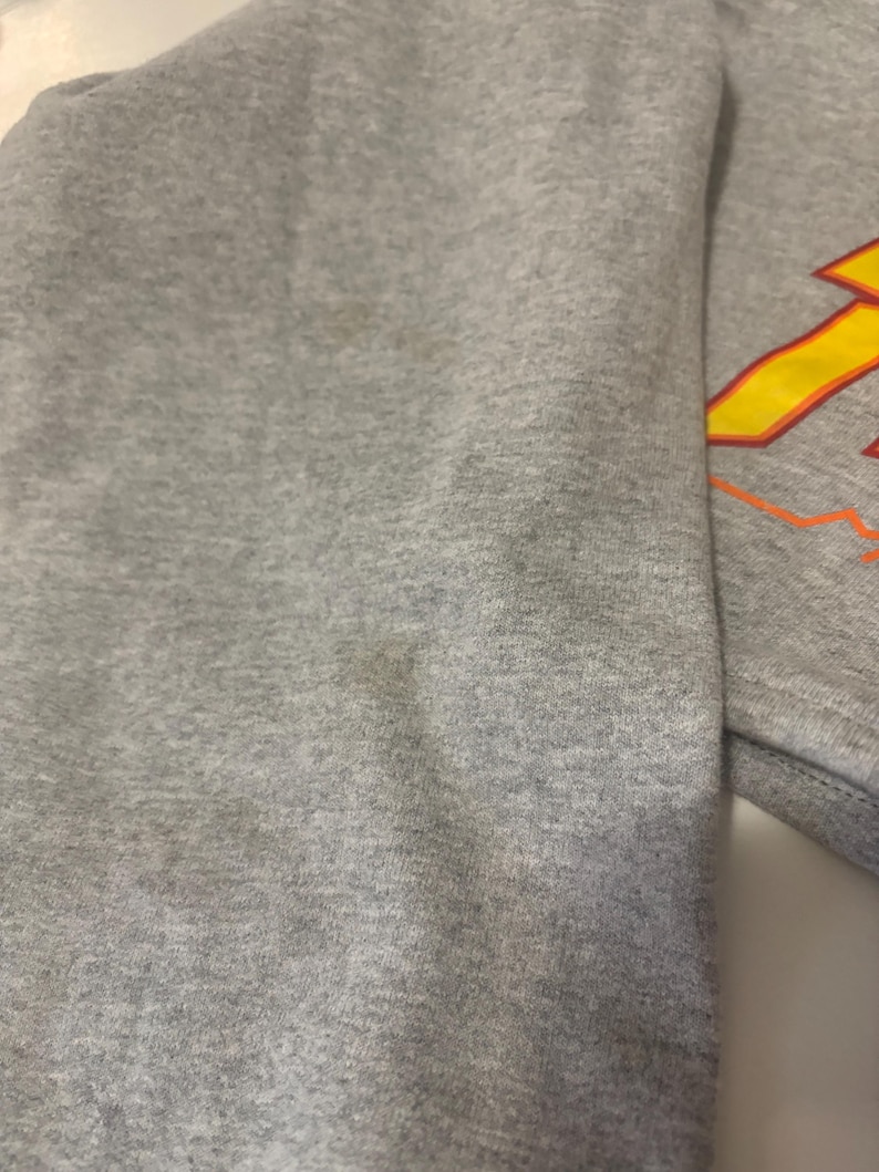 May include: A close-up of a gray sweatshirt. The sleeve is visible, with a design in yellow and orange on the upper right. The fabric has some discoloration.