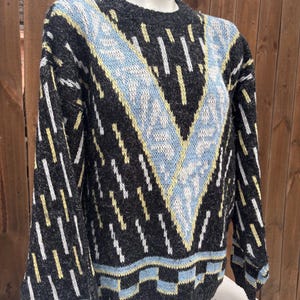 Puede incluir: Un su&eacute;ter gris con un patr&oacute;n geom&eacute;trico en azul claro, amarillo y blanco. El su&eacute;ter presenta un dise&ntilde;o en forma de V en la parte delantera y rayas verticales en las mangas. El cuello y el dobladillo tienen un borde azul claro y negro.