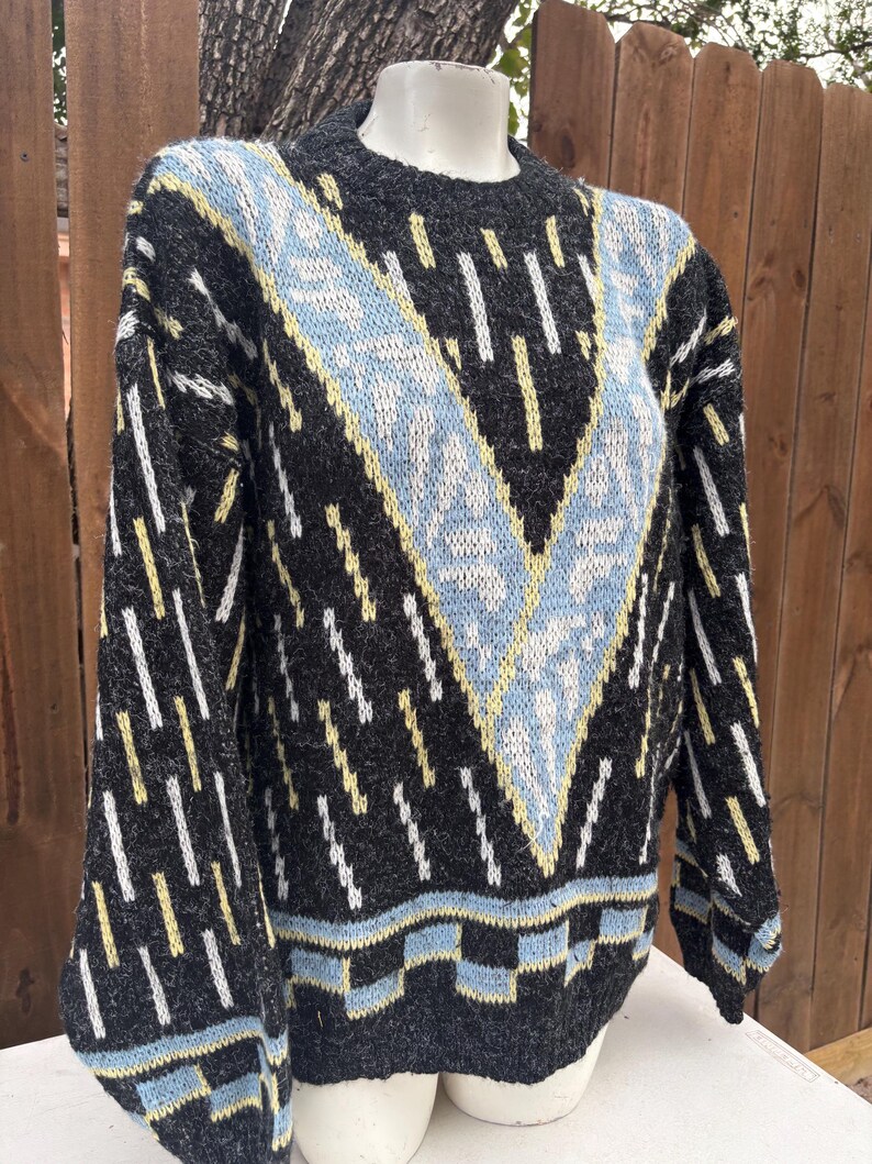 Puede incluir: Un su&eacute;ter gris con un patr&oacute;n geom&eacute;trico en azul claro, amarillo y blanco. El su&eacute;ter presenta un dise&ntilde;o en forma de V en la parte delantera y rayas verticales en las mangas. El cuello y el dobladillo tienen un borde azul claro y negro.