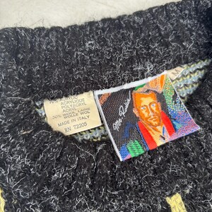Puede incluir: Primer plano de un su&eacute;ter de punto negro y blanco con detalles amarillos y blancos. El su&eacute;ter tiene una etiqueta con el texto "70% ACRYLIC, 30% WOOL" y un retrato de un hombre. El su&eacute;ter est&aacute; hecho en Italia.