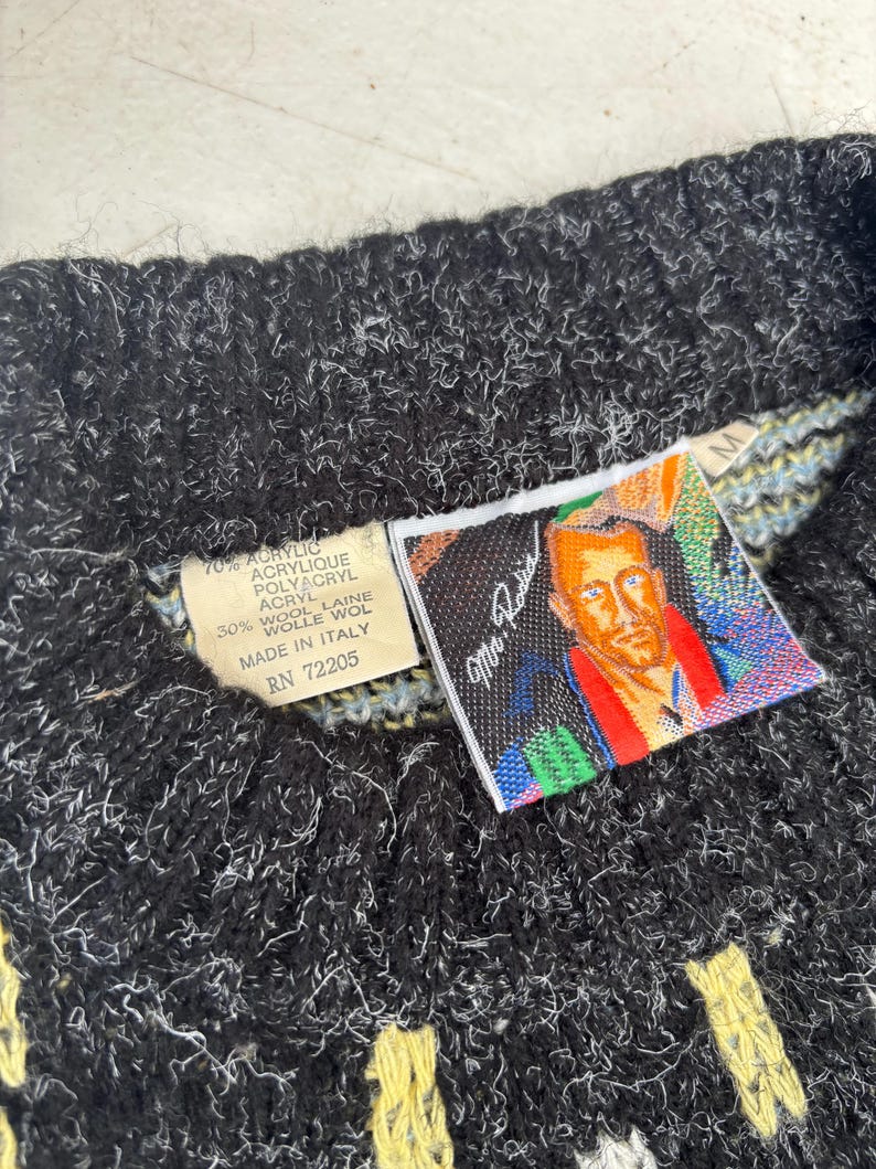 Puede incluir: Primer plano de un su&eacute;ter de punto negro y blanco con detalles amarillos y blancos. El su&eacute;ter tiene una etiqueta con el texto "70% ACRYLIC, 30% WOOL" y un retrato de un hombre. El su&eacute;ter est&aacute; hecho en Italia.