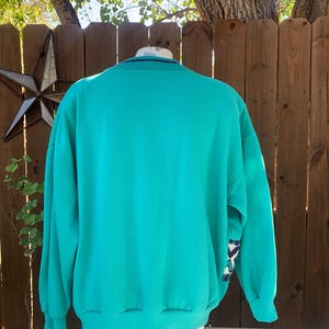 Puede incluir: Sudadera de color turquesa con cuello redondo y mangas largas. Los pu&ntilde;os y el cuello tienen un dise&ntilde;o a rayas. La manga presenta un patr&oacute;n geom&eacute;trico en tonos azules, blancos y negros. La sudadera se exhibe en un maniqu&iacute;.