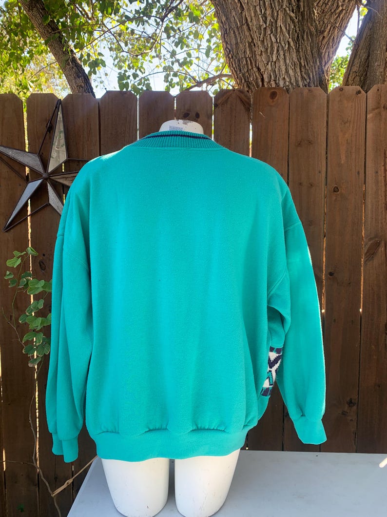 Puede incluir: Sudadera de color turquesa con cuello redondo y mangas largas. Los pu&ntilde;os y el cuello tienen un dise&ntilde;o a rayas. La manga presenta un patr&oacute;n geom&eacute;trico en tonos azules, blancos y negros. La sudadera se exhibe en un maniqu&iacute;.