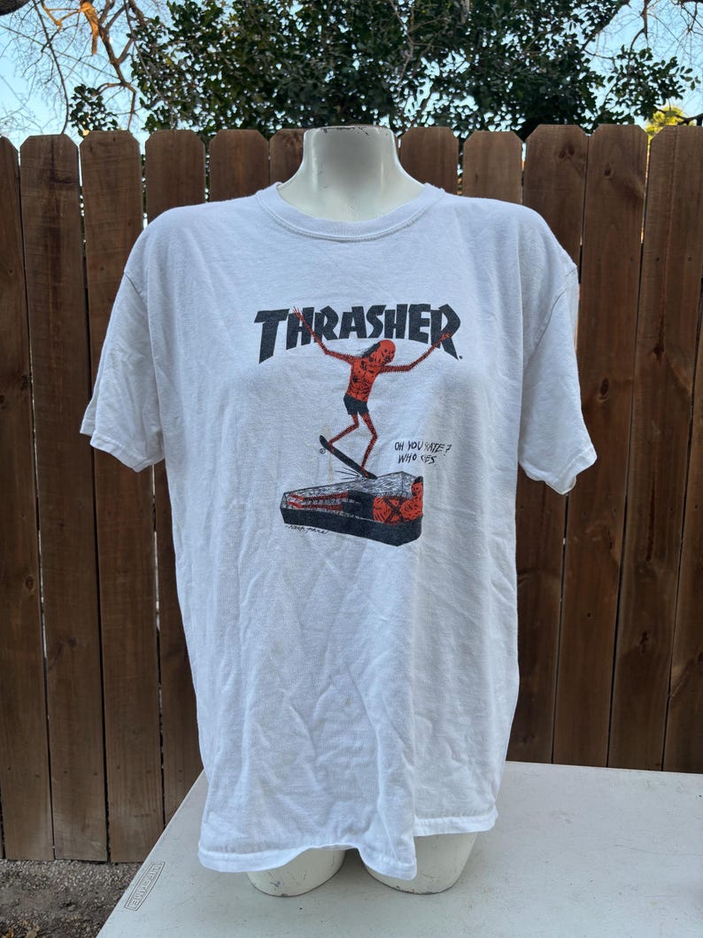 Pu&ograve; includere: T-shirt bianca con il logo Thrasher in rosso e nero. La grafica raffigura una figura di skateboard e il testo "OH YOU SEE? WHO SAYS". La maglietta &egrave; esposta su un manichino.