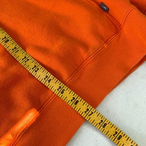 Peut inclure: Un sweat &agrave; capuche orange vif avec un petit logo bleu marine sur le haut de la poitrine gauche. Un m&egrave;tre ruban jaune est pos&eacute; sur le sweat &agrave; capuche, indiquant des mesures de 50,8 &agrave; 76,2 cm.