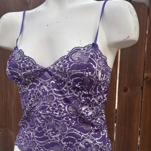 May include: A purple lace camisole with thin spaghetti straps. The camisole features a delicate floral lace pattern and a gathered bust. The garment is displayed on a white mannequin, showcasing the intricate details of the lace.
