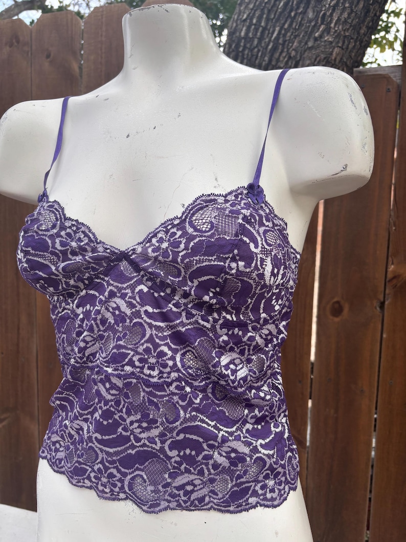 May include: A purple lace camisole with thin spaghetti straps. The camisole features a delicate floral lace pattern and a gathered bust. The garment is displayed on a white mannequin, showcasing the intricate details of the lace.