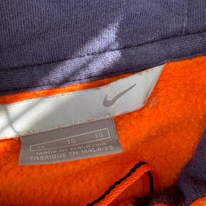 Peut inclure: Gros plan d'un sweat &agrave; capuche orange et bleu marine. L'int&eacute;rieur est orange vif, avec une capuche et des bordures bleu marine. Une &eacute;tiquette avec le logo Nike et le texte "MADE IN MALAYSIA" est visible.