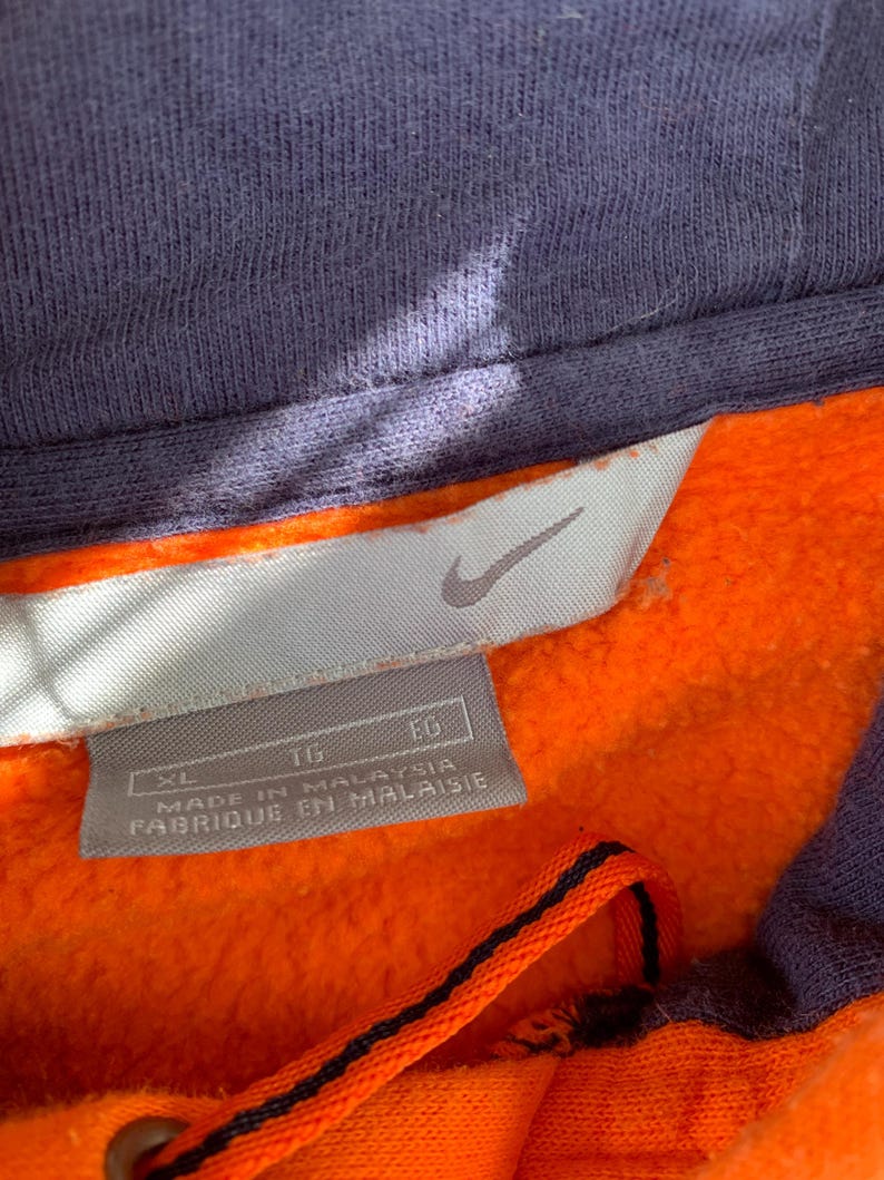 Peut inclure: Gros plan d'un sweat &agrave; capuche orange et bleu marine. L'int&eacute;rieur est orange vif, avec une capuche et des bordures bleu marine. Une &eacute;tiquette avec le logo Nike et le texte "MADE IN MALAYSIA" est visible.
