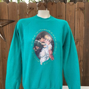 Op de afbeelding: Tealkleurige sweatshirt met een vintage ovale afbeelding van twee kinderen. De afbeelding is omgeven door een bloemenontwerp. De sweatshirt met lange mouwen heeft een ronde hals en wordt op een mannequin getoond.