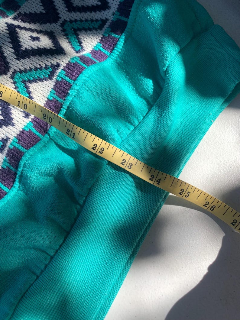 Puede incluir: Primer plano de una manga de sudadera turquesa con un estampado geom&eacute;trico en blanco, turquesa y morado. Una cinta m&eacute;trica amarilla se coloca sobre la manga, mostrando una medida de aproximadamente 56 cm.