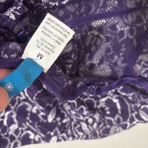 May include: Close-up of a purple lace garment with white floral patterns. The garment has a tag that reads "M" and lists the materials: Nylon 61.4%, Polyester 28.7%, and Elastan 9.9%. The tag also says "Made in Colombia."