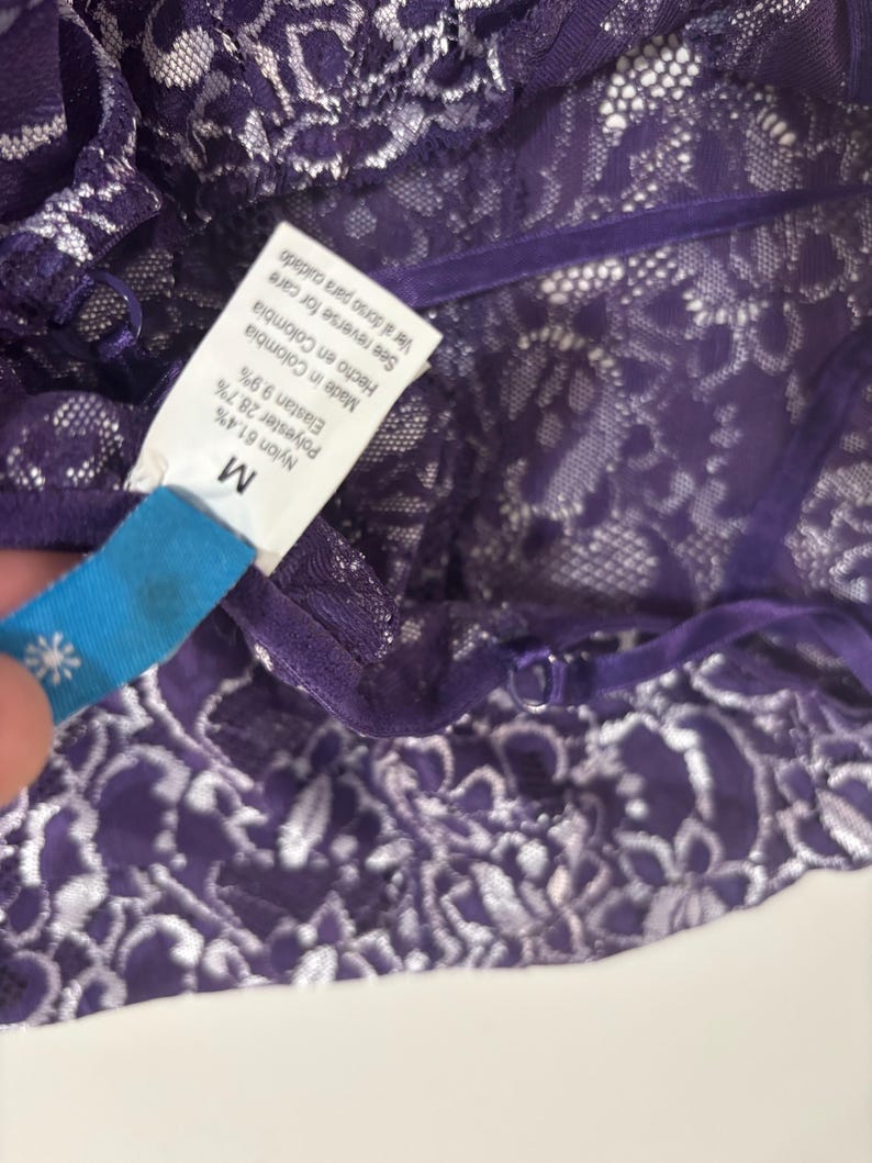May include: Close-up of a purple lace garment with white floral patterns. The garment has a tag that reads "M" and lists the materials: Nylon 61.4%, Polyester 28.7%, and Elastan 9.9%. The tag also says "Made in Colombia."