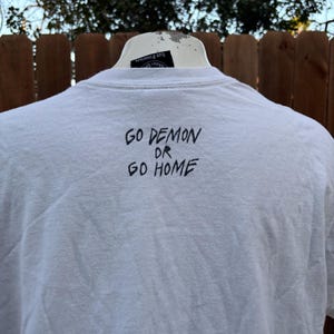 Pu&ograve; includere: T-shirt bianco con la scritta "GO DEMON OR GO HOME" in lettere nere. La maglietta &egrave; esposta su un manichino bianco. Lo sfondo comprende una staccionata in legno e fogliame.