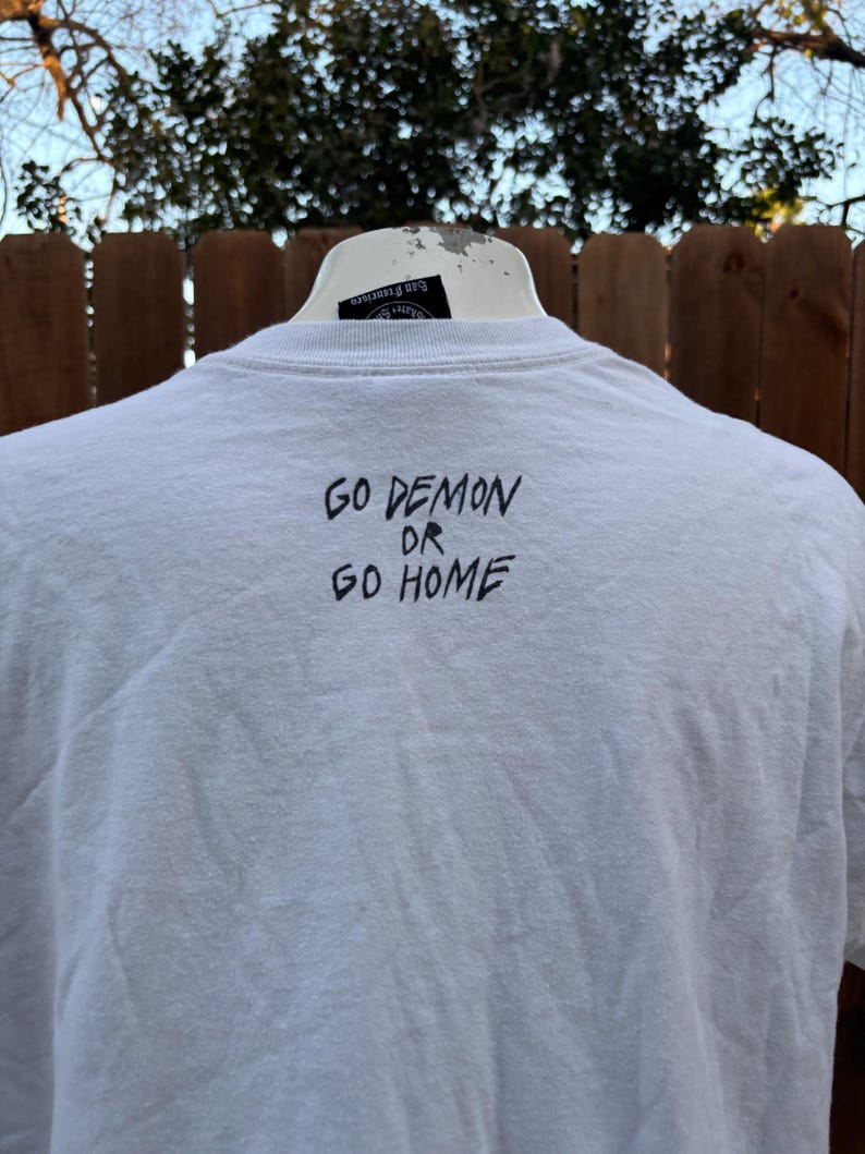 Pu&ograve; includere: T-shirt bianco con la scritta "GO DEMON OR GO HOME" in lettere nere. La maglietta &egrave; esposta su un manichino bianco. Lo sfondo comprende una staccionata in legno e fogliame.