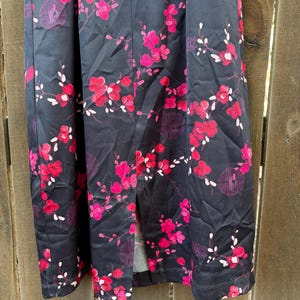 Op de afbeelding: Zwarte stof met een levendig bloemenpatroon met roze en rode bloesems en witte accenten. Het ontwerp bevat takken en cirkelvormige motieven, wat een opvallend visueel contrast cre&euml;ert.