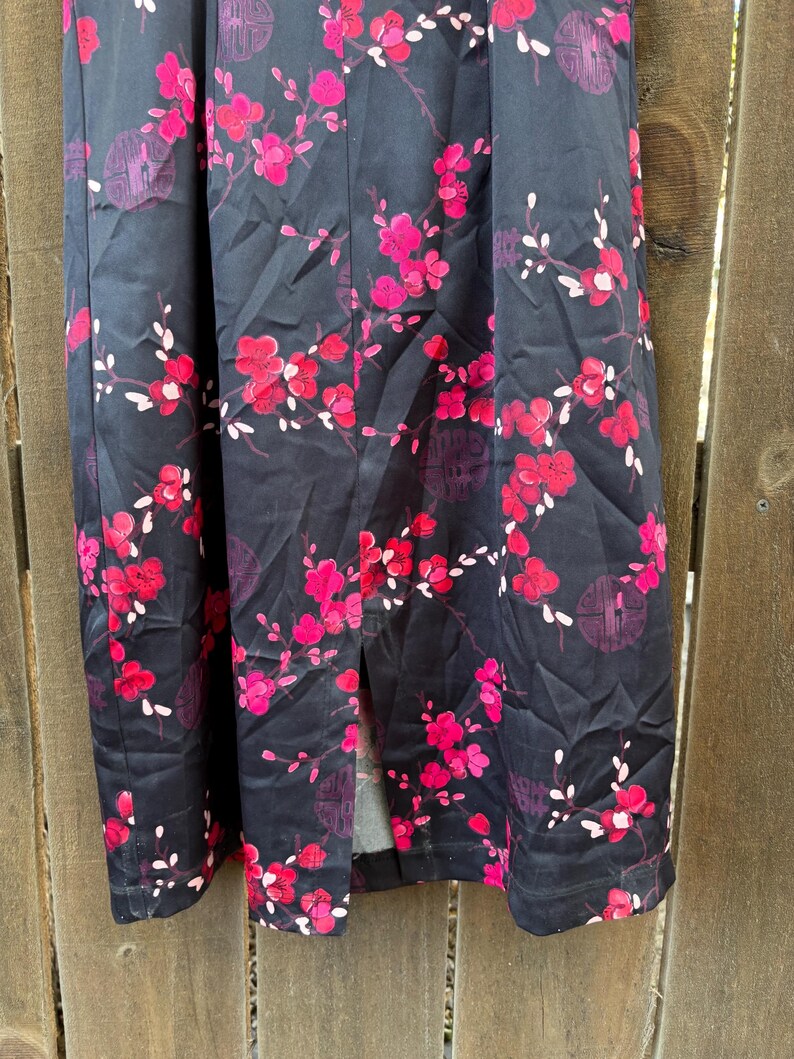 Op de afbeelding: Zwarte stof met een levendig bloemenpatroon met roze en rode bloesems en witte accenten. Het ontwerp bevat takken en cirkelvormige motieven, wat een opvallend visueel contrast cre&euml;ert.