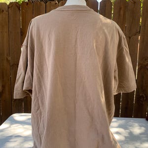K&ouml;nnte beinhalten: Ein hellbraunes T-Shirt mit kurzen &Auml;rmeln, das auf einer Schaufensterpuppe pr&auml;sentiert wird. Das Shirt besteht aus einem weichen, strukturierten Stoff und hat einen Rundhalsausschnitt. Der Hintergrund umfasst einen Holzzaun und &Auml;ste.