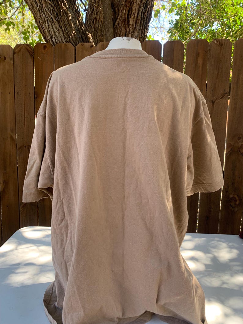 K&ouml;nnte beinhalten: Ein hellbraunes T-Shirt mit kurzen &Auml;rmeln, das auf einer Schaufensterpuppe pr&auml;sentiert wird. Das Shirt besteht aus einem weichen, strukturierten Stoff und hat einen Rundhalsausschnitt. Der Hintergrund umfasst einen Holzzaun und &Auml;ste.