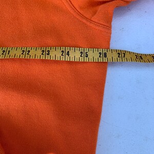 Peut inclure: Gros plan d'un sweat &agrave; capuche orange avec un m&egrave;tre ruban jaune pos&eacute; dessus. Le ruban indique des mesures de 53 &agrave; 71 cm. Le sweat &agrave; capuche est fait d'un tissu doux et textur&eacute;.
