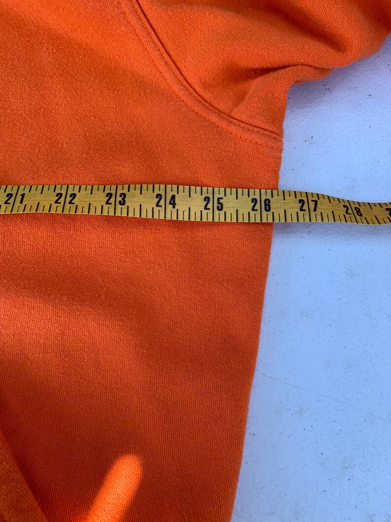 Peut inclure: Gros plan d'un sweat &agrave; capuche orange avec un m&egrave;tre ruban jaune pos&eacute; dessus. Le ruban indique des mesures de 53 &agrave; 71 cm. Le sweat &agrave; capuche est fait d'un tissu doux et textur&eacute;.