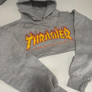 May include: A cropped gray hoodie with a drawstring hood and long sleeves. The front features the word "THRASHER" in yellow, outlined in red, with a flame design. The hoodie is made of a soft, comfortable material.