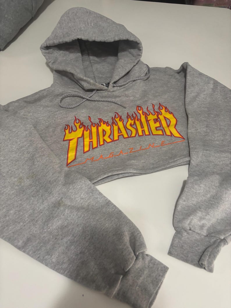 May include: A cropped gray hoodie with a drawstring hood and long sleeves. The front features the word "THRASHER" in yellow, outlined in red, with a flame design. The hoodie is made of a soft, comfortable material.