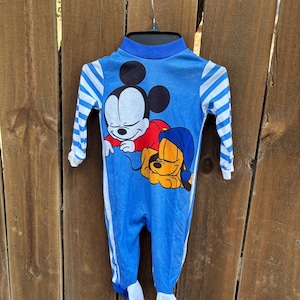 May include: Blue baby sleeper featuring Mickey Mouse and Pluto graphics. The sleeper has long sleeves with white and blue striped cuffs, a blue collar, and white accents down the sides. The feet are white.