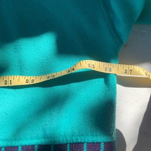 Puede incluir: Un su&eacute;ter color teal con un borde inferior estampado. El borde presenta un dise&ntilde;o geom&eacute;trico en blanco, morado y teal. Una cinta m&eacute;trica amarilla se coloca sobre el su&eacute;ter, mostrando medidas de 53 a 69 cm.